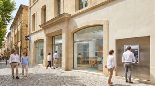 Façade d'une agence bancaire moderne avec des clients marchant sur un trottoir pavé et une personne au distributeur.